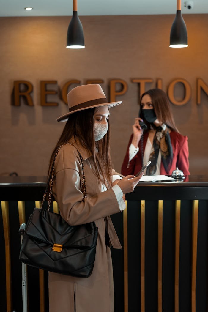 who-we-are Woman in face mask at hotel reception counter checking her phone, symbolizing the new normal.