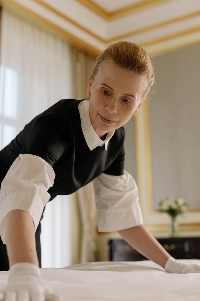 about-us A housekeeper in uniform skillfully arranges bed linen in a luxurious hotel room.